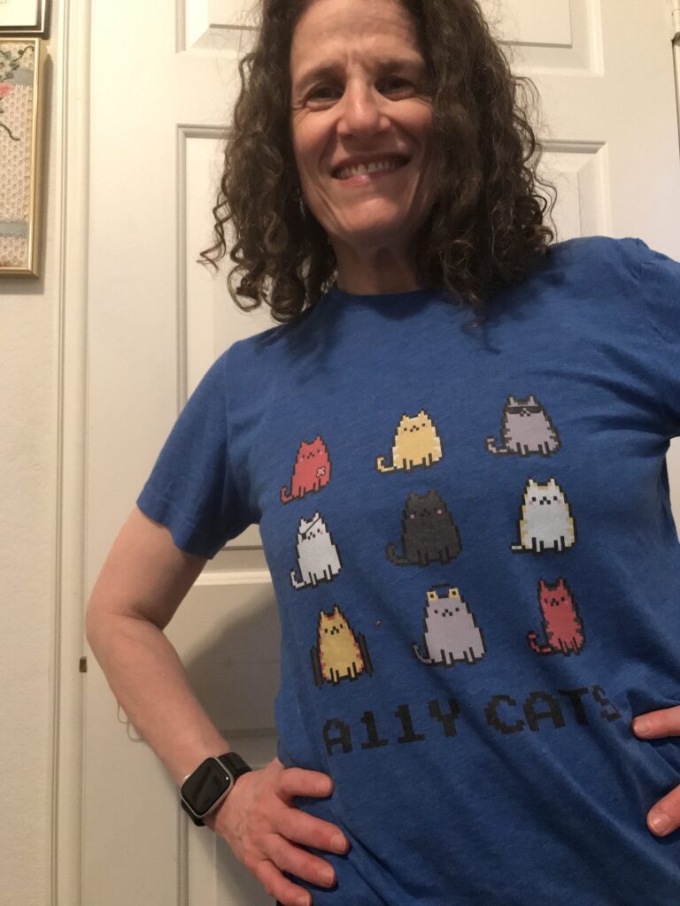 Meryl shows off her a11y cat shirt with 3 x 3 rows of different cats with disabilities