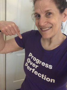 Meryl points to a purple shirt she's wearing that says Progress Over Perfection with an accessibility icon in the O of over